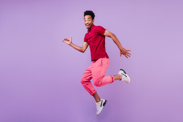 Fototapeta na wymiar Positive carefree african man in sport shoes dancing on purple background. Handsome glad guy in pink pants jumping with smile.