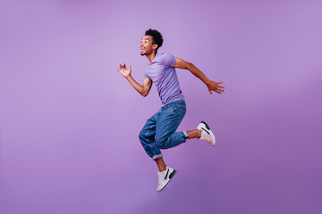 Fototapeta na wymiar Portrait of jumping amazed guy in white sneakers. Indoor shot of dancing stylish male model in purple t-shirt.