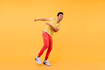 Fototapeta na wymiar Pleased african man with sincere smile dancing on yellow background. Studio photo of enthusiastic black-haired guy wears white sneakers.