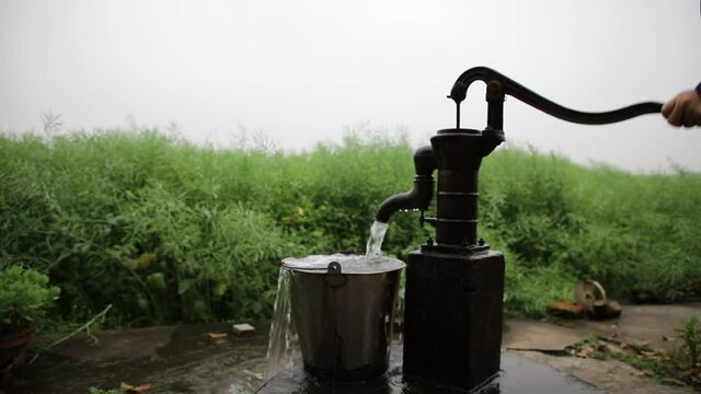 hand pump in front of a countryside village house, providing groundwater as drinking water supply for villagers. Rapeseed weeds field in background