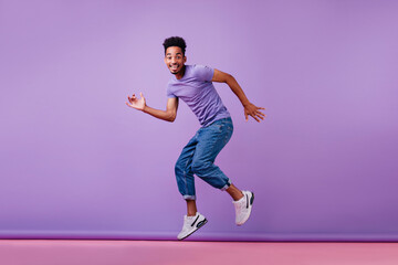 Good-humoured male model dancing in new white sneakers. Studio shot of ecstatic african guy in jeans.