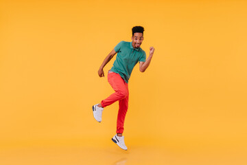 Fototapeta na wymiar Full-length shot of inspired black guy dancing in red pants. Indoor photo of carefree african man enjoying photoshoot on yellow background.