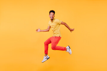Fototapeta na wymiar Dreamy male model with brown skin fooling around during photoshoot. Indoor shot of african guy in bright clothes jumping on yellow background.