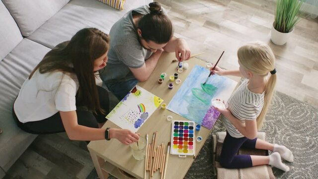 Young parents, mom and dad, are drawing funny pictures with their daughter, using watercolors, having nice communication together, enjoy common hobby, Top view, Slow motion.