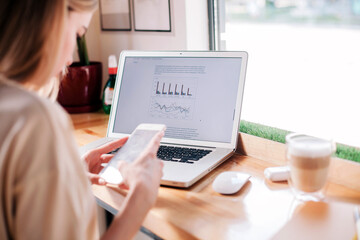 Young girl with phone in hand works behind a laptop in a coffee shop and drinks a cappuccino