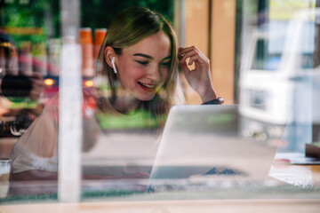 Fototapeta premium Girl in coffee shop window works on laptop sipping cappuccino