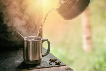 hot tea is poured from a teapot into a metal mug with steam on a background of grass.