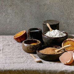 Ingredients for home baking. Flour, eggs, cane sugar, milk in different ceramic bowls, blood oranges and spices on linen table cloth. Grey texture background