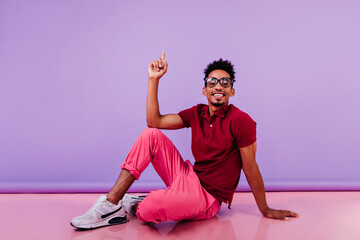 Carefree stylish guy sitting on the floor. Confident male model in pink pants looking to camera with happy face expression.