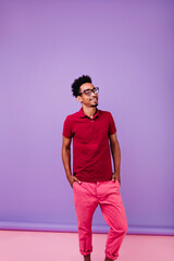 African male model in good mood enjoying photoshoot on violet background. Short-haired optimistic man posing with hands in pockets.