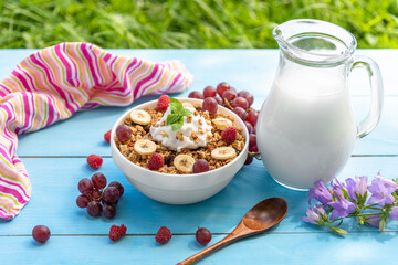 Breakfast cereals with milk and raspberries, grapes, banana in the open air on a light blue table and on a background of green grass.