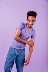Laughing lovable man in jeans posing in studio. Indoor photo of african male model with trendy haircut standing on purple background.