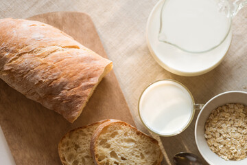 Light rustic breakfast - rustic bread with cut pieces, a cup of oatmeal, a jug and a glass of milk on a linen napkin on the table by the window, top view, morning light
