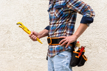 A construction worker in a blue checked shirt with tools in his belt holds a yellow wrench in his hands.