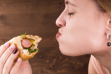 Young attractive woman eating hot dog on background of brown wooden wall, close-up