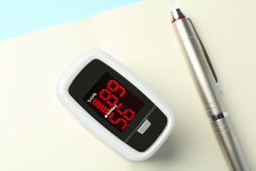 Modern fingertip pulse oximeter, pen and open notebook on light blue background, flat lay