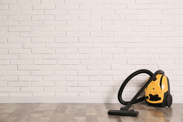 Modern yellow vacuum cleaner on wooden floor near white brick wall, space for text