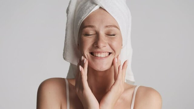 Young attractive woman with towel on head happily touch her cleaned face and rejoicing on camera over gray background