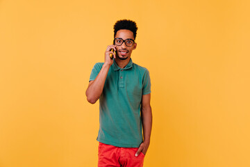 Surprised black male model talking on phone. Studio shot of stylish african guy in glasses posing on yellow background with smartphone.