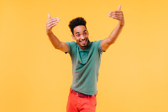 Emotional stylish man in green t-shirt having fun in studio. Carefree black guy waving hands on yellow background.