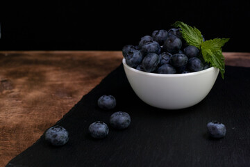 Blueberries with mint leaves in a small white bowl. On a black granite board. Yummy.
