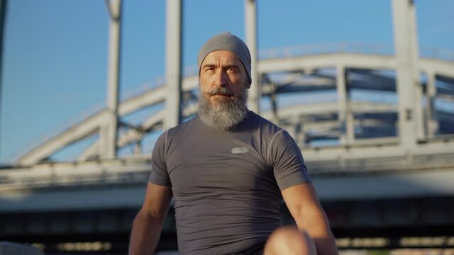 Zoom in portrait of senior athlete with grey beard looking at camera while doing warm up exercises outside