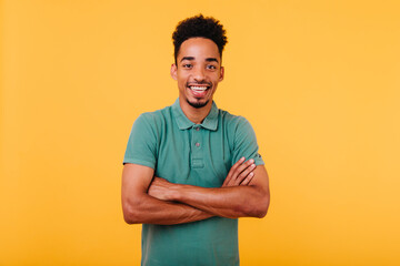 Laughing african guy with trendy haircut posing on bright yellow background with arms crossed. Studio shot of black man smiling to camera during photoshoot.