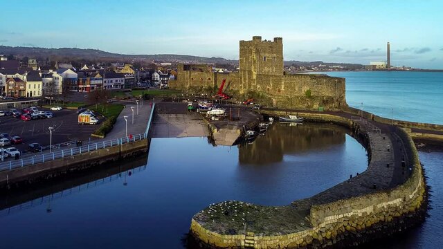 Medieval Norman Castle and harbor in Carrickfergus near Belfast in sunset light. Aerial 4K flyby/approaching/revealing video with water reflection, marina, boat ramp, breakwater and town 