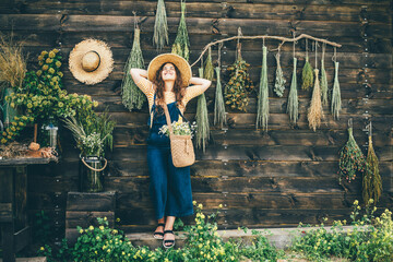 attractive girl wearing straw hat and blue denim dungarees hangs bouquets of aromatic wildflowers...