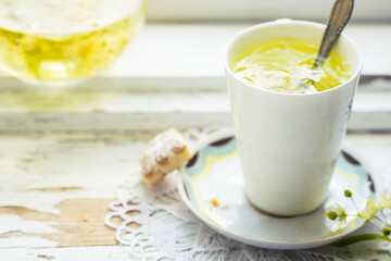 Obraz na płótnie Canvas Linden flowers tea in a white cup with silver spoon on a white napkin, on a wooden background and cookie nearby, yellow drink of linden flower on a vintage background, healthcare and healthy eating co