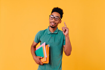 Smart curly guy with books posing with smile. Indoor shot of interested african student isolated on yellow background.