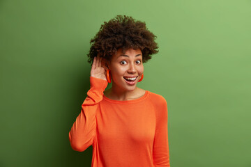 Good looking cheerful woman with curly hair keeps hand near ear as tries to overhear interesting conversation, listens carefully coworkers who talk privately, being glad and curious, stands indoor