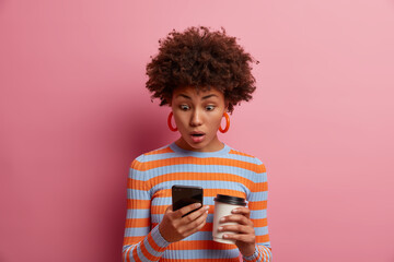 Astonished surprised curly young woman stares at smart phone display, sees something amazing online, reads disturbing insulting message, drinks takeout coffee, poses against pink background.
