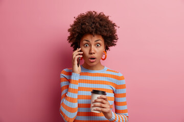 Impressed shocked woman hears fresh outstanding news, talks via phone, opens mouth from surprise, holds takeaway coffee, expresses amazement, wears casual striped jumper, isolated on pink wall