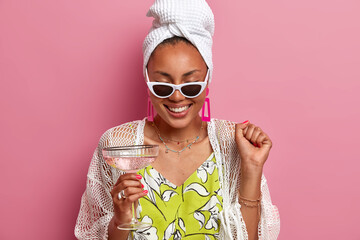 Cheerful housewife has fun on pajama party, raises clenched fist, celebrates special event, drinks cocktail, wears bath towel on head, sunglasses, giggles positively, isolated over pink background