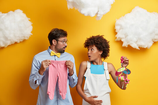 Shocked woman and man stare at each other with embarrassement, come to training lesson for future parents, pose with mobile and babys clothes, are not prepared for parenthood. Child birth concept