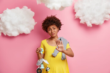Expectant mother makes refusal gesture, keeps palm forward camera in rejection, has pregnant round tummy, poses with baby stuff against pink wall with white clouds. Parenting and motherhood.