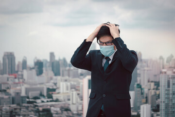 Young Asian businessman in suit wearing face mask with skyscraper city outdoor, stressed frustrated man putting his head in his hand thinking about his job, feeling disappointed or tired.