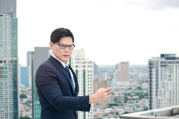 Young Asian businessman in suit using smart phone on the rooftop with skyscraper city view