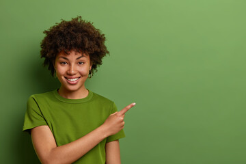 Friendly looking glad female shop assistant happy to help customers, shows way and demonstrates discounts in shop, points fore finger aside on empty space over green background. Advertising concept