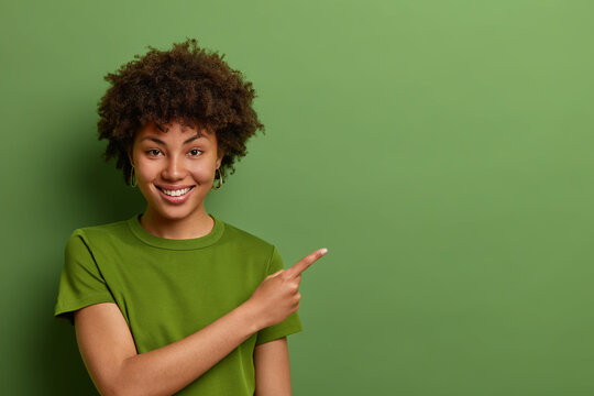 Friendly looking glad female shop assistant happy to help customers, shows way and demonstrates discounts in shop, points fore finger aside on empty space over green background. Advertising concept
