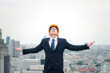 Cheerful and happy achievement successful young Asian businessman engineer in suit wearing safety helmet raising his arm up to celebrate success goal on the rooftop with skyscraper city view