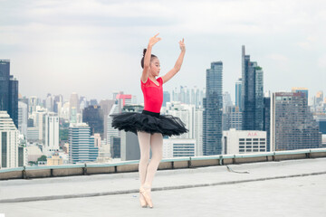 Fototapeta na wymiar Asian ballerina dancer girl practicing ballet dancing on rooftop with skyscraper city view, adorable child dancing in ballet