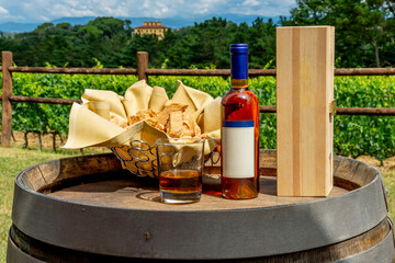 A bottle of vin santo with its wooden box and a tray of cantuccini resting on top of a wooden barrel with the Tuscan countryside in the background, Pisa, Italy