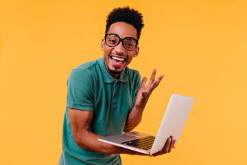 Handsome male freelancer in glasses smiling on yellow background. Ecstatic african student holding laptop and expressing happiness.