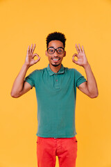Naklejka premium Inspired young man posing with surprised smile and making okay sign. Studio portrait of enthusiastic guy in colorful attire and glasses.
