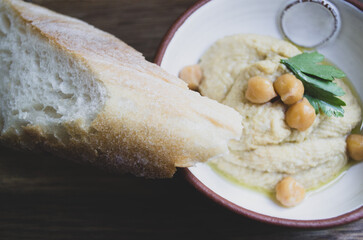 Hummus decorated with parsley and chickpea peas in a ceramic bowl with a piece of bread on a wooden background