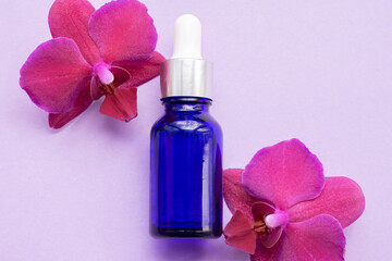 Fototapeta na wymiar Blue glass bottle filled by essence or serum with orchid extract on purple background with bright blossoming orchids phalaenopsis. Eco cosmetic concept