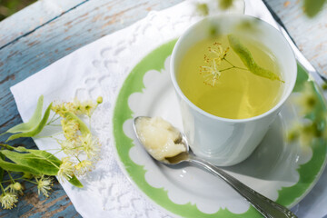 Linden flowers tea in a white cup on a white with green ornament plate with spoon and honey on a blue wooden background and white napkin outdoors, yellow drink of linden flower on a vintage background