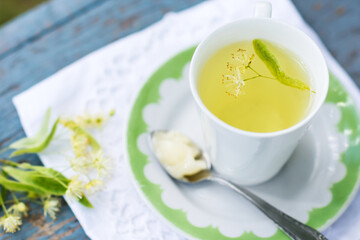 Linden flowers tea in a white cup on a white with green ornament plate with spoon and honey on a blue wooden background and white napkin outdoors, yellow drink of linden flower on a vintage background